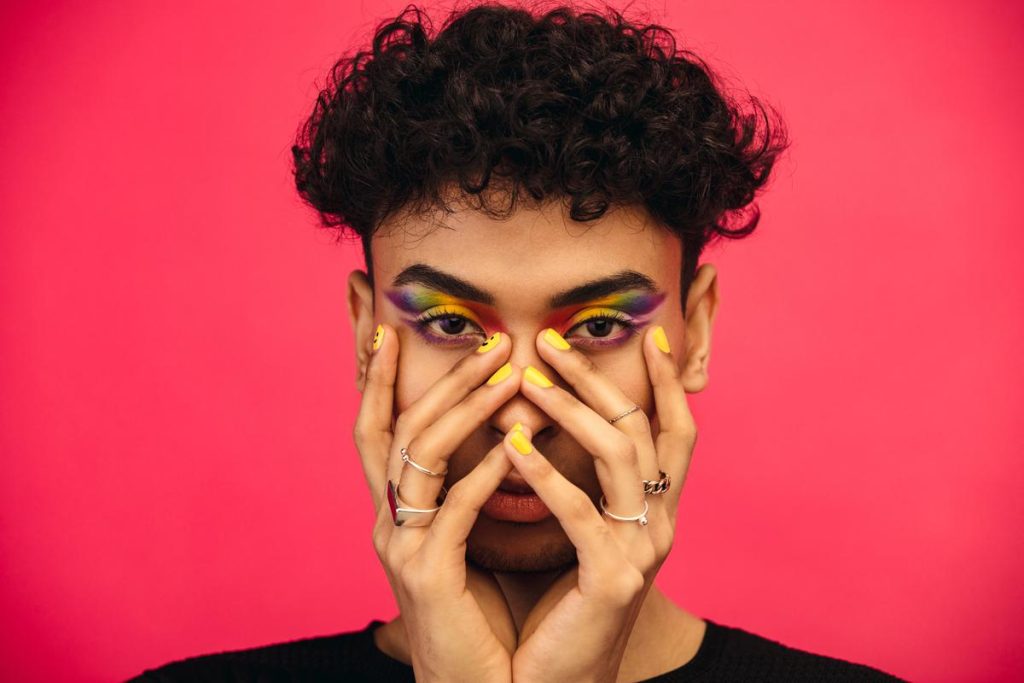 an individual with short curly hair, yellow painted fingernails, fingers adorned with rings, and rainbow eyeshadow. they are shown with their hands on their face against a red background.