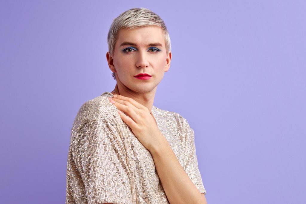 an individual with short blonde hair wearing a sparkly champagne colored shirt shown holding their shoulder against a lavender background