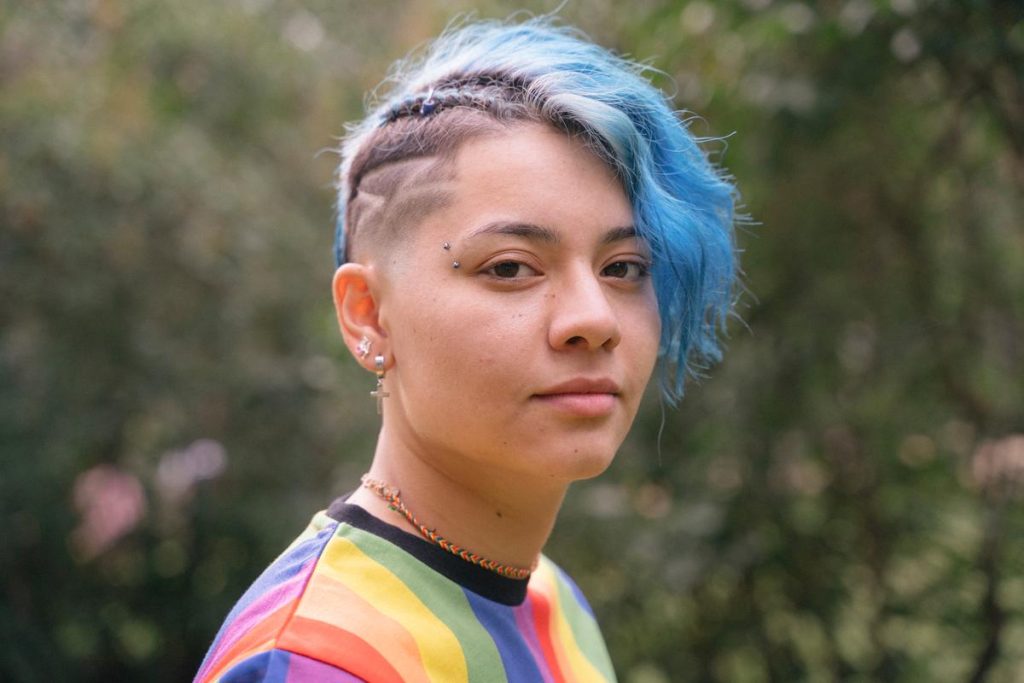 an individual with blue hair is shown standing outdoors wearing a rainbow shirt.